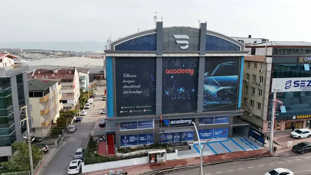 Binaya daha yakın çekilen havadan karede giriş katı boyunca sıralanan eğitim afişleri seçiliyor: Renkli Kaplama, PDR, Polisaj, PPF, Cam Filmi ve 'Araç Bakımında Usta Eğitimi'. Girişin üstünde 'Ustalığa giden yol burada başlar' yazıyor.