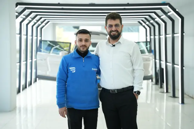 Bir katılımcı ile beyaz gömlekli kişi, aynı koridorun LED kemerleri önünde omuz omuza poz veriyor; arkada bir araç duruyor. Bu kare, sertifika takdimlerinin çekildiği yerde alınmış.