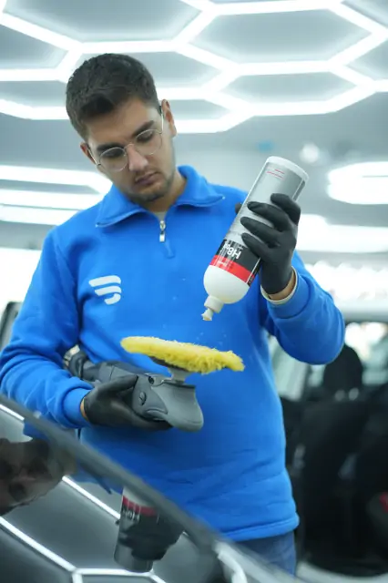 Makineye takılı sarı yün pedin üzerine şişeden pasta veriliyor; diğer el makineyi gövdesinden tutuyor. Pasta panele değil pede ve az verilir; fazlası çalışırken etrafa savrulur, kenarlarda birikir ve silinirken iz bırakır.