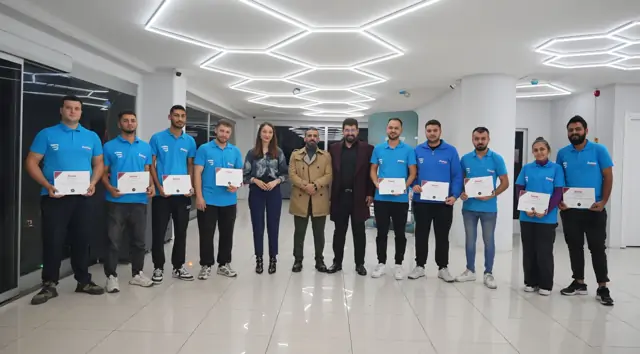 Salonda toplanan grubun büyük bölümü elinde eğitim sertifikasıyla poz veriyor; camlardan dışarısı karanlık, kare gün bitiminde çekilmiş. Grupta kadın katılımcılar da var.