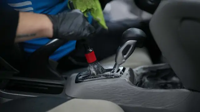 Detay fırçası köpükle vites konsolunun dokulu yüzeyinde çalışıyor, diğer elde yeşil mikrofiber bez bekliyor. Dar ve desenli iç yüzeylerde ürün doğrudan panele değil fırçaya verilir; böylece düğme ve yuvalara sıvı kaçmaz.