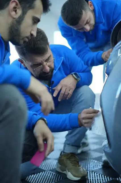 Üç kişi aracın arka kanadının önüne çökmüş; biri ıslak filmi panel hattı boyunca bıçakla kesiyor, diğerinin elinde pembe ragle duruyor. Panel üzerinde kesim yapılırken bıçağın açısı ve baskısı belirleyicidir; amaç yalnızca filmi kesmek, boyaya hiç dokunmamaktır.