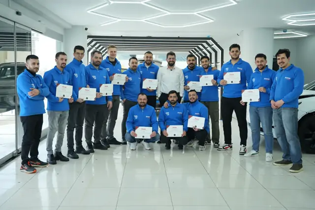 Kalabalık bir grup, LED kemerli koridorun önünde toplanmış; öndeki üç kişi çömelmiş, çoğunun elinde eğitim sertifikası var. Arkada beyaz bir araç duruyor.