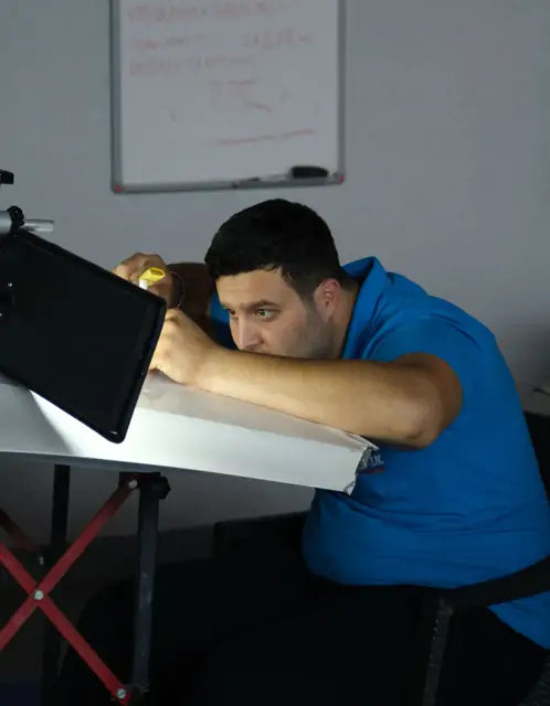 Katlanır masaya alınmış beyaz panelin üzerine siyah çerçeveli bir yansıma panosu eğik biçimde konumlandırılmış; bir kişi göz hizasında yüzeye bakarak yansımayı takip ediyor, arkadaki beyaz tahtada kırmızı kalemle alınmış notlar duruyor. Boyasız göçük düzeltmede hasar doğrudan değil, ışığın yüzeyde kırılma biçiminden okunur.