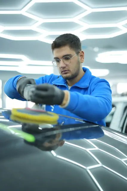 İki elle kavranan makine siyah kaputta sarı pedle çalışıyor; panel kenarına yeşil koruma çekilmiş, altıgen aydınlatma yüzeyde yansıyor. Tek elle bastırılan makine yüzeyde sıçrar ve düzelttiğinden fazla iz bırakır.