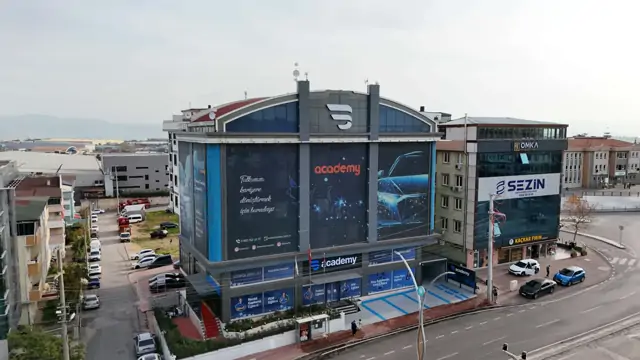 Havadan çekilen karede kemer çatılı bina köşe konumuyla, çevresindeki sokak ve mavi çizgili otoparkıyla birlikte görünüyor. Cephedeki yazı okunuyor: 'Tutkunuzu kariyere dönüştürmek için buradayız...'