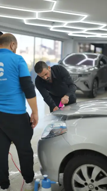 Gümüş bir aracın ön bölümüne serilen ıslak film pembe ragleyle bastırılıyor; yerdeki el pompası ve zemine akan sabunlu su uygulamanın ıslak yapıldığını gösteriyor. Ragle çekildikçe film ile boya arasındaki su dışarı atılır; su tamamen çıkmadan yapıştırıcı boyayı tutmaz.