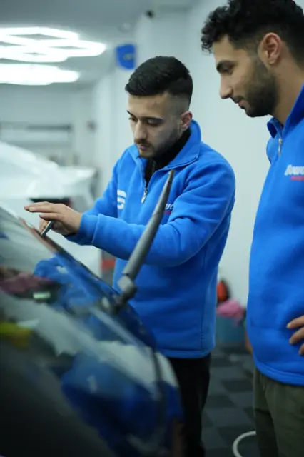 Silecek kaldırılmış ön camda bir kişi parmak uçlarıyla filmin kenarını bastırıyor, yanındaki kişi işlemi izliyor. Camın alt kenarı sileceklerin sürekli süpürdüğü bölgedir; buradaki film ucu tam yapışmazsa kısa sürede kalkar.