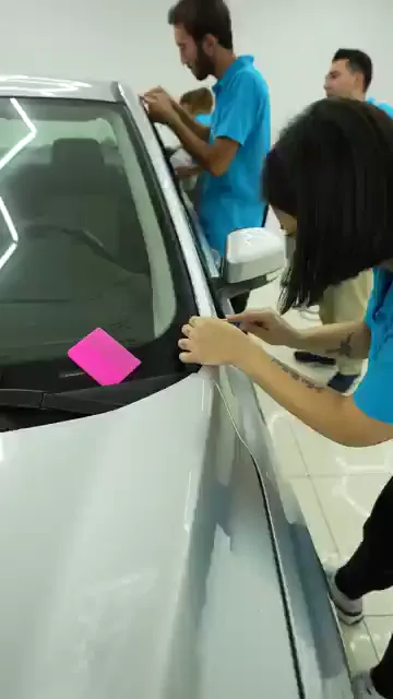 Yakın planda ön direğin kapı fitili görünüyor. Katılımcı bir eliyle lastik fitili kenara alıyor, diğer eliyle ince uçlu bir aletle filmin kenarını fitilin altına itiyor. Direğin üst ucunda ikinci bir kişi aynı fitili yukarıdan tutuyor; pembe ragle o sırada camın üzerinde bekletiliyor.