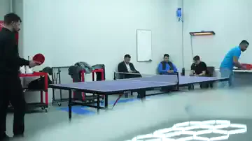 Uzun masanın bir ucunda siyah kıyafetli bir kişi servis atıyor, rakibi kadrajın dışında kalıyor. Masanın arkasındaki sıralarda oturan birkaç kişi maçı takip ediyor; duvarda beyaz tahta ve kırmızı yangın dolabı görünüyor. Oyun sürerken oturanlar yerlerinden kalkmıyor, oda ikili bir kullanımda kalıyor.