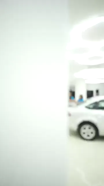 Kamera odanın girişinden ilerliyor; altıgen aydınlatmanın altında beyaz bir sedanın arka bölümünde üç kişi eğilmiş halde çalışıyor. Yerde iki basınçlı pompa ve serili hortum var, aracın çevresinde her yönden dönülebilecek kadar boşluk bırakılmış. Ekip aynı panelin üst kenarına birlikte uzanıyor.