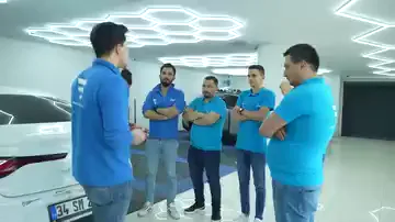 Şapkalı bir kişi, karşısında yarım halka kuran mavi tişörtlü gruba dönük konuşuyor. Halkanın ortasında beyaz bir araç duruyor, tavandaki altıgen aydınlatma parlak zeminde ikinci kez görünüyor. Gruptakiler araca değil konuşana bakıyor; anlatım henüz araç üzerine geçmemiş.