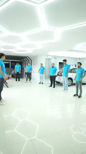 Beyaz zeminli geniş bölümde grup, karşılarındaki kişiye dönük olarak sıraya geçiyor. Ardından herkes kollarını göğüs hizasında öne ve yanlara doğru açıp kapatıyor. Arkada park hâlinde beyaz bir araç ve bir vantilatör duruyor; hareket, günlük çalışma alanının ortasında yapılıyor.