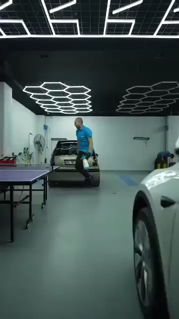 Park hâlindeki araçların arasına açılmış masa tenisi masası, çalışma alanının tam ortasında duruyor. Mavi tişörtlü bir kişi masanın çevresinde dolaşıp elindeki şişeyi tezgâha bırakıyor; masanın üzerinde raketler hazır bekliyor. Duvarda yangın dolabı, tavanda altıgen aydınlatma var: oyun için ayrı bir oda açılmamış.