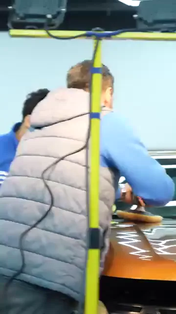 Turuncu kaputun üzerine pasta zikzak çizgiler hâlinde bırakılmış; bir kişi sarı pedli makineyi bu çizgilerin üzerine oturtup çalıştırıyor. Yanında iki kişi kaputun başında durmuş, makinenin izlediği yolu takip ediyor. Tavandaki altıgen aydınlatma boyada olduğu gibi yansıdığı için pasonun geçtiği yer ile geçmediği yer aynı karede ayırt ediliyor.