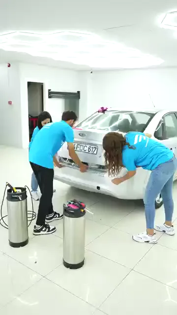 Beyaz bir sedanın arkasında üç katılımcı aynı anda çalışıyor: biri bagaj kapağının üst kenarına yatırılan filmi pembe ragleyle bastırıyor, diğerleri tamponun iki yanındaki kenarları tutuyor. Yerde iki basınçlı pompa ve hortumu duruyor, panel ıslak. Film, ıslatılmış yüzeyde konumlandırıldıktan sonra kenarlara doğru sıyrılıyor.
