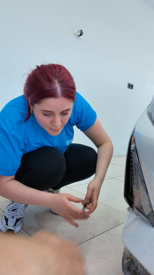 PPF kaplama eğitiminde kadın kursiyerimiz ön tampon ve far çevresine film uyguluyor; far ve ızgara kenarlarında fazla filmi kesip kenarları kapatıyor.