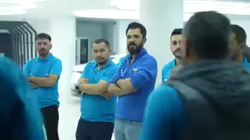 Aynı mavi tişörtü giyen bir grup, kollarını kavuşturmuş hâlde yarım daire oluşturmuş duruyor. Kameraya sırtı dönük bir kişi onlara doğru konuşuyor; arkadaki bölümde beyaz bir araç ve dolap sırası görünüyor. Konuşma bir masa ya da tahta başında değil, doğrudan çalışma alanının içinde geçiyor.