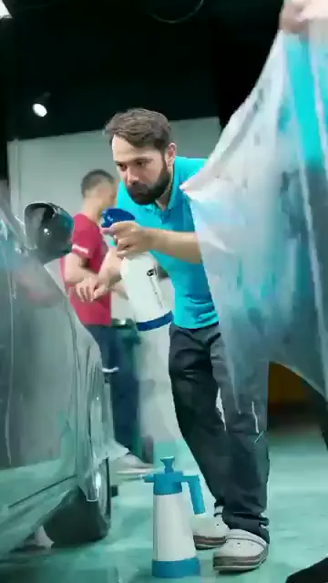 Geniş bir şeffaf film parçası aracın yanına açılıyor; yerde sabunlu su spreyi duruyor ve katılımcılar uygulamayı birlikte izliyor.

Eğitmen filmi çekerken ölçülü olunmasını hatırlatıyor: sert çekilen film bir anda boşalıp elde kalabiliyor. Bu ürünü doğru çekebilenin diğer filmlerde zorlanmayacağını da ekliyor.

Son karede pembe ragle ile filmin kenarına birlikte bakılıyor.