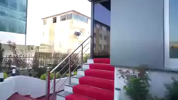 Kamera dışarıdaki merdivenden başlayıp cam cepheli girişe doğru ilerliyor; basamaklar ve giriş holü kırmızı halıyla kaplanmış. İçeride büyük bir ekran yanıyor, cam yüzeyler karşıdaki sokağı ve binaları yansıtıyor. Gökyüzü kararmaya başlamış, çekim tek bir kesintisiz hareketle yapılmış.