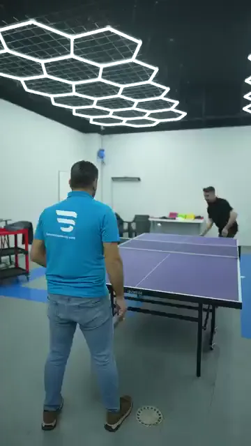 Tesisin dinlenme bölümünde iki kişi masa tenisi oynuyor; masa mavi, tavan altıgen aydınlatmayla kaplı. Servis atılıyor, top masanın üzerinden geçiyor ve ralli birkaç vuruş sürüyor. Masanın ucunda renkli raketler, duvar kenarında sandalyeler duruyor.