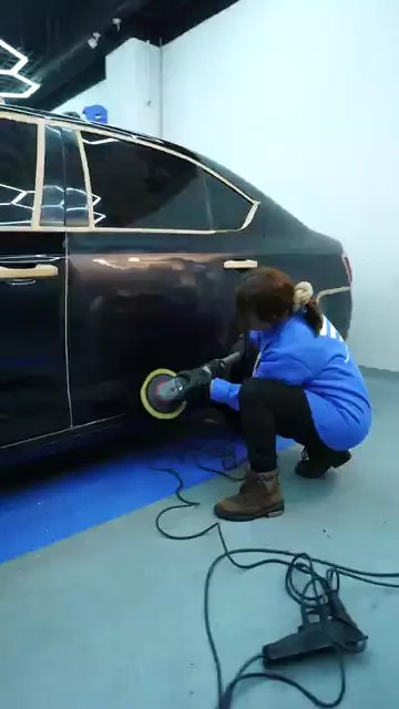Cam çerçevesi ve kapı kolu maskeleme bandıyla kapatılmış koyu bir aracın yanında, at kuyruklu bir kadın çömelip sarı pedli makineyi arka kapının alt bölgesinde gezdiriyor. Kablo yerde serbest bırakılmış, tavandaki altıgen aydınlatma boyada boydan boya yansıyor. Bu hizaya ulaşmak için ayakta durmak yerine zemine iniliyor.