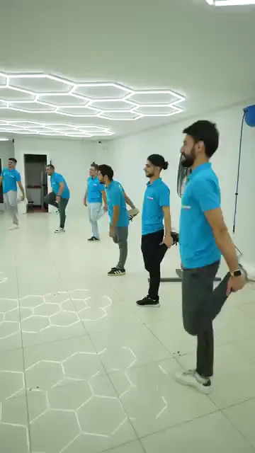 Sıra hâlinde dizilen grup bir ayağını arkaya çekip bileğinden tutuyor ve tek ayak üzerinde dengede kalmaya çalışıyor. Herkes aynı yöne dönük, hareket aynı anda yapılıyor. Tavandaki altıgen aydınlatma parlak zeminde ikinci bir desen oluşturuyor.