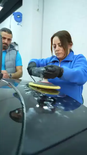Koyu renk aracın kaputuna pasta damlaları bırakılmış; mavi sweatshirtlü, eldivenli bir kadın sarı pedli makineyi yüzeye oturtup kısa pasolarla ilerletiyor. Arkasında duran bir kişi makineye dokunmadan yalnızca izliyor. Pasolar kaputun ön kenarı boyunca ilerleyip aynı bölgeye birkaç kez dönüyor.