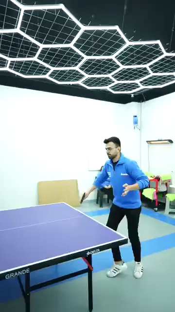 Mavi sweatshirtlü bir oyuncu masanın bu ucunda gövdesini çevirip topa vuruyor, rakibi kadraja girmiyor. Arka duvardaki sıralarda oturan iki kişi oyunu izliyor, tavanı altıgen aydınlatma kaplıyor. Kareler arasında yalnızca oyuncunun duruşu değişiyor; ayakları aynı noktada kalıyor.