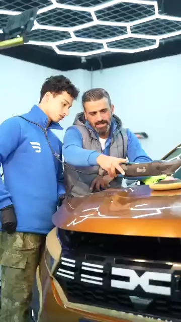 Aynı makinenin üzerinde iki el var: mavi sweatshirtlü genç katılımcı gövdeyi tutuyor, yanındaki kişi elini makinenin ön kısmına koyup yönü birlikte veriyor. Turuncu kaputta pasta çizgileri duruyor ve makine bu çizgilerin üzerinden geçiyor. Kareler boyunca ikinci el makineden ayrılmıyor, yalnızca ilerleme hızı değişiyor.