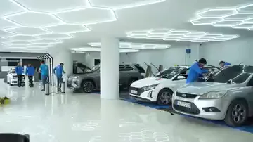 Beyaz zeminli geniş bir salonda birden fazla araç yan yana duruyor; tavanı baştan sona altıgen LED hatları kaplıyor. Mavi üniformalı ekipler ayrı araçların çevresinde çalışıyor, zeminde bekleyen ped ve kablolar görülüyor. Her aracın dört yanında dönüp çalışacak kadar boşluk bırakılmış.