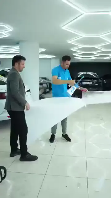 Beyaz bir sedanın bagaj kapağına şeffaf film yatırılmış; bir katılımcı ragleyle ortadan kenara doğru suyu sıyırırken, diğeri filmin stop lambası tarafındaki kenarını gergin tutuyor. Panel ıslak, filmin fazlası tamponun üzerine sarkıyor.