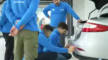Arka tampona film uygulanıyor: ayakta duran bir kişi filmi üst kenardan gergin tutarken, çömelen iki kişi stop lambası çevresindeki kenarı pembe ragleyle bastırıyor. Çevrelerinde mavi sweatshirtlü diğer katılımcılar durup işlemi izliyor; hepsi aynı panele bakıyor.