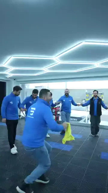 Ekip el ele tutuşup bir sıra oluşturmuş; sıranın önündeki kişi elindeki sarı mendille dizlerini kırıp yere yakın figürler yapıyor. Arkadaki sıra tempoyu bozmadan bekliyor, kimileri gülüyor. Zemin kare karo, arka planda camlı cephe ve servis alanı görünüyor.