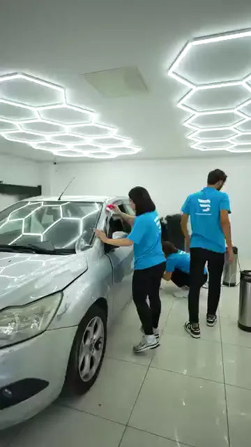Gümüş renkli bir aracın ön direğine şeffaf film yatırılmış. Katılımcı filmin kenarını bir eliyle gergin tutarken diğer eliyle pembe ragleyi direk boyunca aşağı doğru bastırıyor; filmin artan kısmı ön camın üzerine sarkıyor. Yüzey ıslak, hemen yanında basınçlı pompa duruyor ve aynı aracın alt kenarıyla ikinci bir katılımcı ilgileniyor.