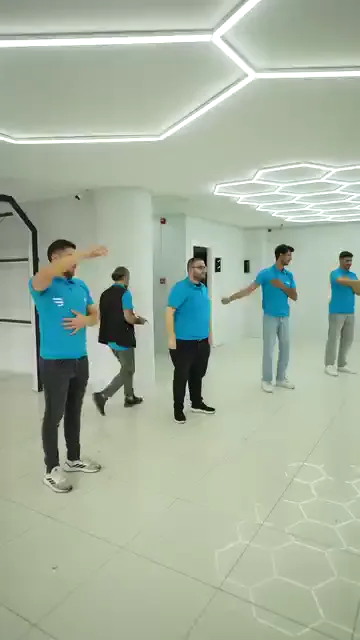 Boş ve aydınlık bir salonda mavi tişörtlü bir grup kollarını iki yana açarak aralarındaki mesafeyi ayarlıyor. Ardından herkes aynı anda kolunu yukarı kaldırıp hareketi tekrarlıyor. Alanda araç ya da tezgâh yok; zemin bu iş için tamamen boşaltılmış.