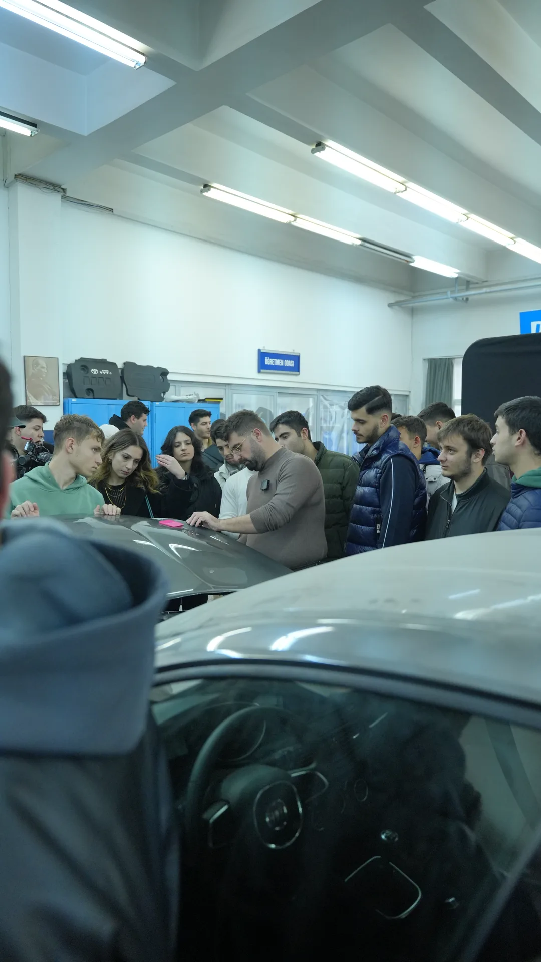 Bir üniversite etkinliğinde öğrenciler aracın çevresinde toplanmış, eğitmen kaputta film uygulamasını gösteriyor